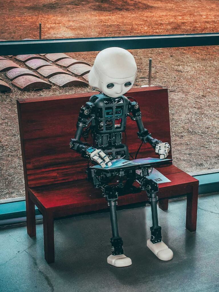 A photo of a robot setting at a bench reading on a tablet