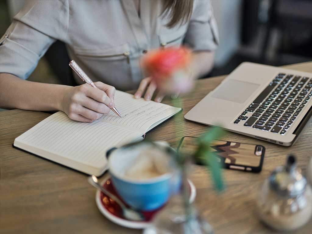 a close up of a lady's hand writing in a notebook with laptop and mobile on the desk